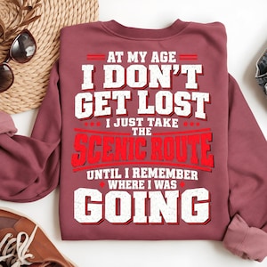 May include: A maroon crewneck sweatshirt featuring white and red text that reads: AT MY AGE I DON'T GET LOST I JUST TAKE THE SCENIC ROUTE UNTIL I REMEMBER WHERE I WAS GOING. The lettering has a distressed, weathered texture throughout.