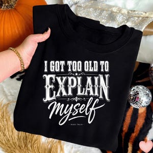 Può includere: Una felpa nera a girocollo con testo grafico bianco che recita I GOT TOO OLD TO EXPLAIN MYSELF con il nome MARK TWAIN stampato in lettere più piccole sottostanti. Il design utilizza un mix di caratteri in grassetto e in corsivo.