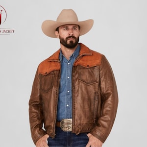 May include: A brown leather jacket with a contrasting orange yoke and collar. The jacket has two chest pockets and a belt buckle. The model is wearing a cowboy hat and a denim shirt. The Washington Jacket logo is in the upper left corner.