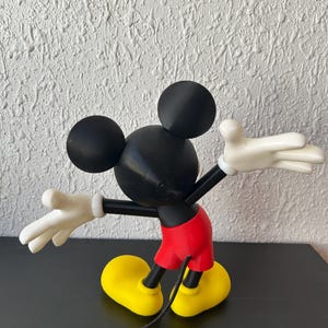 May include: A Mickey Mouse figurine shown from the back, featuring a black head with round ears, a black torso, bright red shorts, white gloves with four fingers, and large yellow shoes. A thin black tail extends downward from the red shorts.