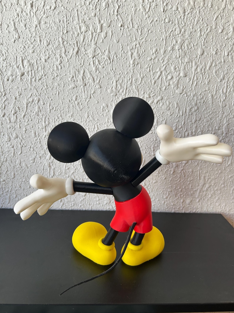 May include: A Mickey Mouse figurine shown from the back, featuring a black head with round ears, a black torso, bright red shorts, white gloves with four fingers, and large yellow shoes. A thin black tail extends downward from the red shorts.