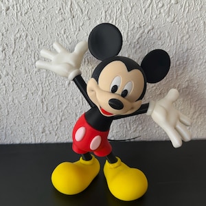 May include: A Mickey Mouse figurine featuring a black head with large round ears, a tan face, and a black nose. The character wears red shorts with two white oval buttons, white gloves, and large yellow shoes, posed with arms outstretched.