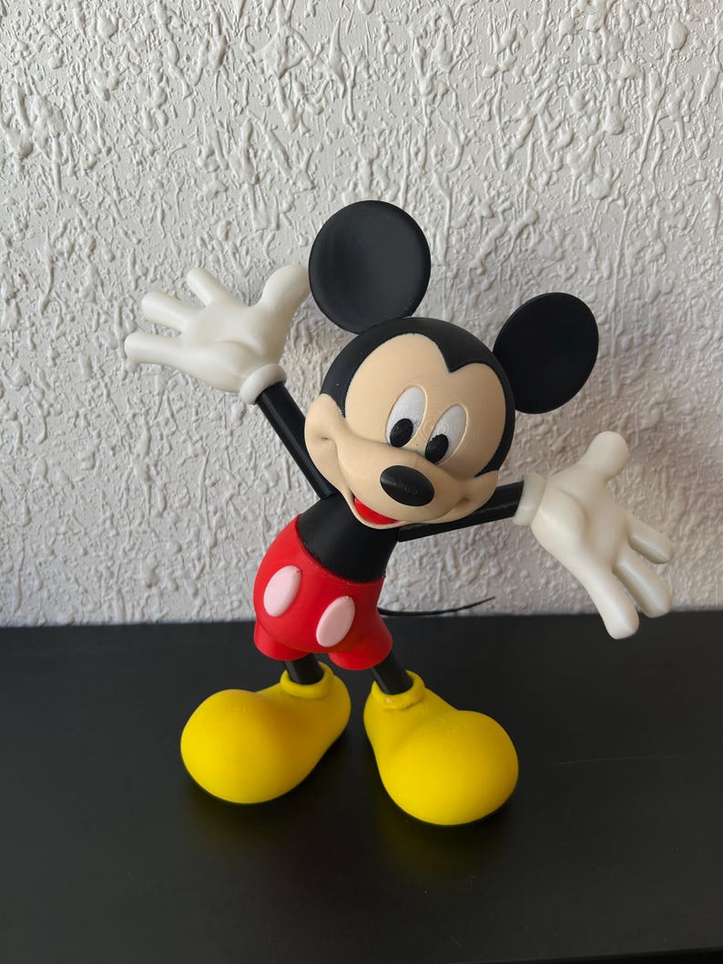 May include: A Mickey Mouse figurine featuring a black head with large round ears, a tan face, and a black nose. The character wears red shorts with two white oval buttons, white gloves, and large yellow shoes, posed with arms outstretched.