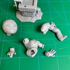 Puede incluir: Un kit de modelo impreso en 3D de resina gris que presenta una estructura de pozo cuadrada con textura de ladrillo y varias piezas desmontadas de una figura de superh&eacute;roe, incluyendo una cabeza con patr&oacute;n de red, un torso, una pierna y dos manos.