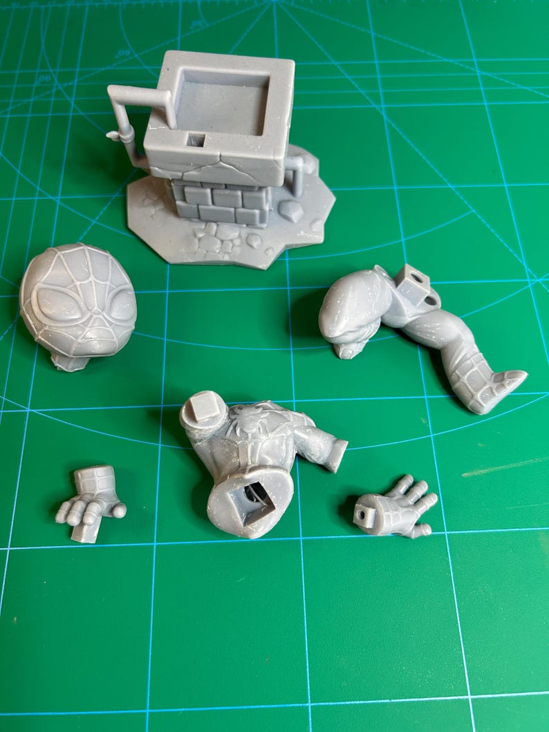 Puede incluir: Un kit de modelo impreso en 3D de resina gris que presenta una estructura de pozo cuadrada con textura de ladrillo y varias piezas desmontadas de una figura de superh&eacute;roe, incluyendo una cabeza con patr&oacute;n de red, un torso, una pierna y dos manos.