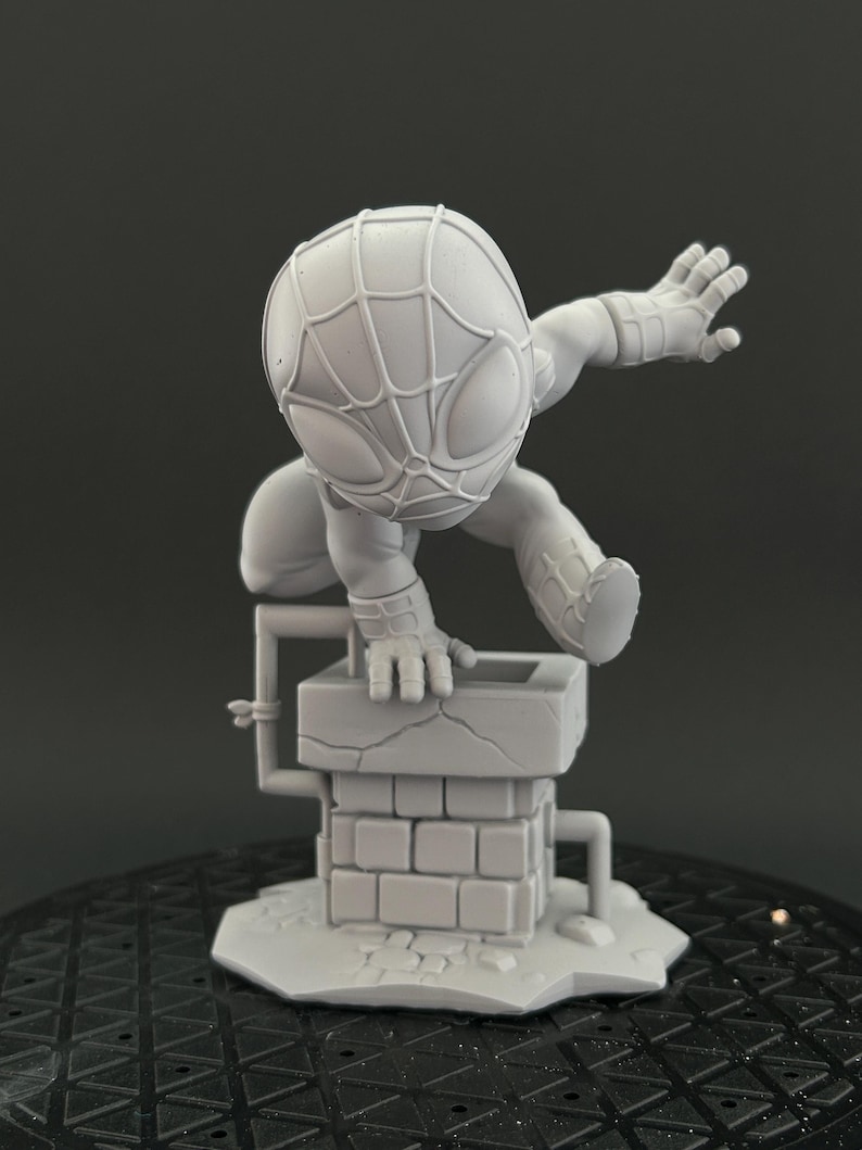Puede incluir: Una figura de resina de color gris claro sin pintar de un superh&eacute;roe estilizado con cabeza grande y patr&oacute;n de m&aacute;scara de telara&ntilde;a. La figura est&aacute; agachada en una pose din&aacute;mica sobre una peque&ntilde;a estructura de chimenea de ladrillo con tuber&iacute;as.