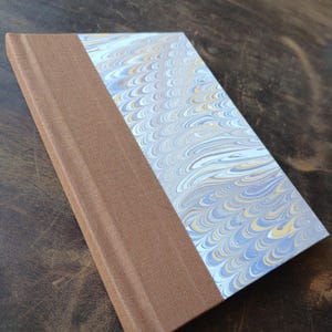 May include: A hardcover journal featuring a brown fabric spine and a marbled paper cover with a repeating wave pattern in shades of light blue, white, and yellow. A small red ribbon bookmark extends from the bottom edge of the book binding.