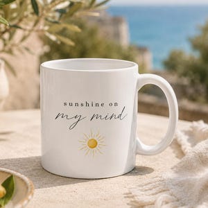 Könnte beinhalten: Weiße Keramik-Tasse mit dem Text "sunshine on my mind" in schwarzer Schrift und einer gelben Sonnenillustration. Die Tasse hat einen gebogenen Henkel und steht auf einer hellen Oberfläche.
