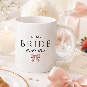 Könnte beinhalten: Ein weißer Keramikbecher mit dem schwarzen Text "IN MY BRIDE era" und einer kleinen rosa Schleifenillustration, die unter den Wörtern zentriert ist. Der Becher hat einen Standardhenkel und eine glatte Oberfläche für den täglichen Gebrauch.