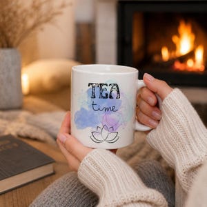 Könnte beinhalten: Weiße Keramiktasse mit den Worten "TEA time" in schwarzer Schrift und einem Lotusblumen-Design. Die Tasse hat ein Aquarelldesign in Blau- und Lilatönen. Die Tasse wird mit beiden Händen gehalten.