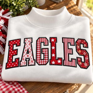 May include: A white mock neck sweatshirt featuring the word EAGLES in large, block-style letters. Each letter is filled with a different red and white pattern, including polka dots, floral prints, gingham checks, stars, and vertical stripes.