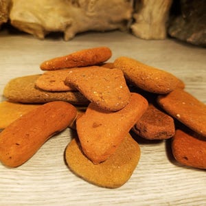 Pode incluir: Uma coleção de pedras de argila planas e lisas na cor terracota com uma textura porosa e terrosa. As pedras variam em forma, indo de arredondadas a alongadas, e exibem tons de castanho avermelhado, laranja queimado e ocre suave.