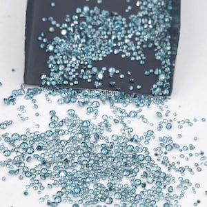 May include: A large collection of small, round, faceted blue gemstones scattered across a white surface, with many more contained within a dark gray metal scoop featuring the white text Alakhdiam printed on its surface.