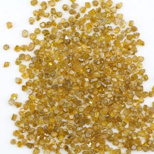 May include: A large collection of small, raw, unpolished yellow diamonds. Each individual stone features irregular, multifaceted shapes with a translucent, golden-yellow hue. The diamonds are scattered across the frame, displaying natural textures and varying levels of clarity and brilliance in their uncut, rough state.