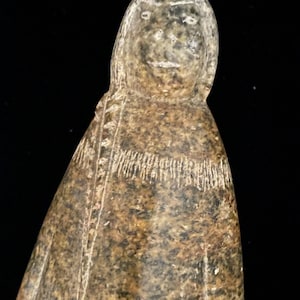 Puede incluir: Una figura de piedra tallada que representa a una persona con cabello largo y una superficie texturizada con motas marrones y grises. La figura viste una prenda con un patrón grabado horizontal en el pecho y líneas verticales a los lados del cuerpo.