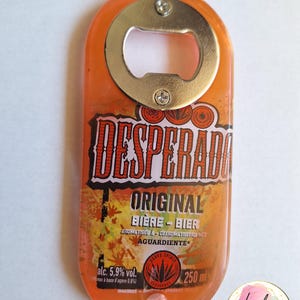 Pu&ograve; includere: Apribottiglie in resina arancione con un apribottiglie in metallo. L'apribottiglie presenta il testo "DESPERADO ORIGINAL BIERE-BIER" e "alc. 5,9% vol.". L'apribottiglie &egrave; lungo 10 cm e largo 4 cm.