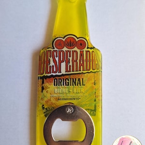 Pu&ograve; includere: Un apribottiglie giallo a forma di bottiglia con la scritta "DESPERADOS ORIGINAL BIERE-BIER" e "AGUARDIENTE". L'apribottiglie ha un apribottiglie in metallo argentato. Il design include elementi grafici arancioni e marroni.