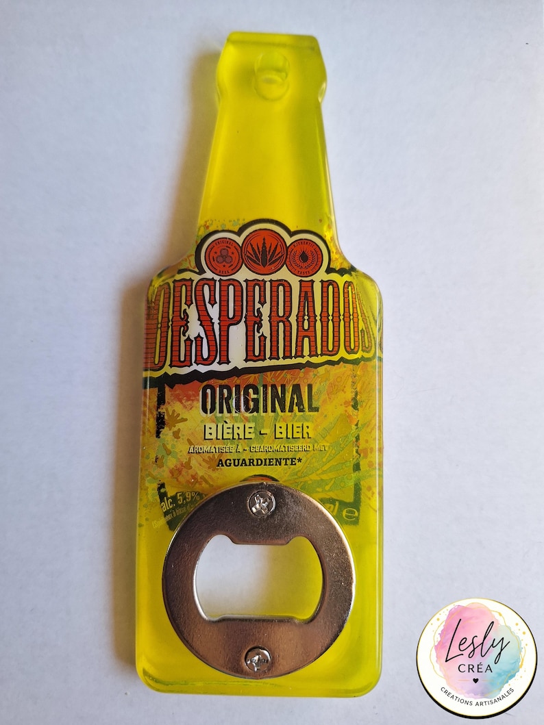 Pu&ograve; includere: Un apribottiglie giallo a forma di bottiglia con la scritta "DESPERADOS ORIGINAL BIERE-BIER" e "AGUARDIENTE". L'apribottiglie ha un apribottiglie in metallo argentato. Il design include elementi grafici arancioni e marroni.