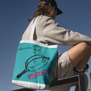 May include: A turquoise tote bag featuring a black and white illustration of a frying pan and spoon. The bag has the text "#WINNING" in pink. It has white straps and a white border.