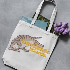 May include: Cream tote bag with a cartoon cat illustration and the Spanish text "Comiendo Mierda" in yellow. The bag contains books and purple tulips.