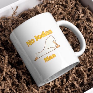 Peut inclure: Tasse en céramique blanche avec l'inscription "No Jodas Mas" en jaune, accompagnée d'une illustration de canard. La tasse est dans une boîte blanche remplie de papier froissé brun.