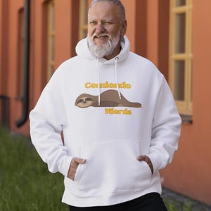 May include: White hoodie with a front pocket and a drawstring hood. The hoodie features a graphic of a sloth and the text "Comiendo Mierda" in yellow. The sloth is brown and the text is in a playful font.