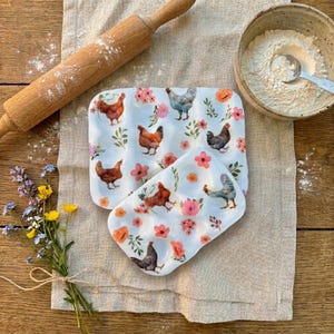 May include: Two white ceramic egg trays with a floral and chicken design. A wooden rolling pin and a bowl of flour are also present. The trays are decorated with pink and orange flowers and various chicken illustrations.