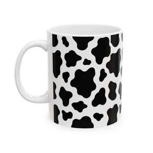 May include: A white ceramic mug featuring a repeating black cow print pattern across the exterior surface. The mug includes a curved handle on the left side and a cylindrical body, showcasing a high-contrast black and white spotted design.