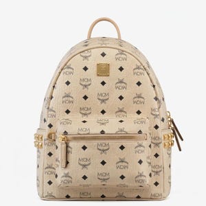 May include: Beige backpack with a repeating black MCM logo pattern and black diamond shapes. The backpack has a top handle, a front pocket with a gold zipper, and gold-colored studs on the sides. The MCM logo is also visible on a gold-colored plaque.