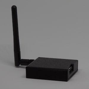 Op de afbeelding: Een zwarte rechthoekige draadloze netwerkadapter met een enkele verticale antenne aan de zijkant. Het apparaat heeft een getextureerde matte afwerking en een zichtbare micro-USB-poort op het zijpaneel, ontworpen voor connectiviteit en gegevensoverdracht.