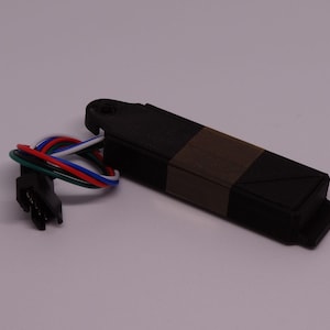Könnte beinhalten: Ein rechteckiges, schwarzes elektronisches Sensormodul mit einem grauen Mittelstreifen und einem kleinen Montageloch an einem Ende. Ein kurzer Kabelbaum mit vier farbigen Drähten—rot, grün, blau und weiß—ragt seitlich heraus und endet in einem schwarzen Stecker.