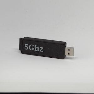May include: A black rectangular USB Wi-Fi adapter featuring the text 5Ghz printed in white on its side. The device has a standard metallic USB connector extending from one end, designed for wireless network connectivity in electronic devices.