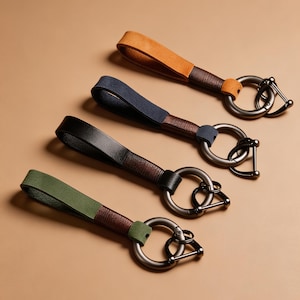 May include: Four keychains arranged in a diagonal row, each featuring a looped strap in orange, navy blue, black, or olive green. The straps are secured with dark brown leather wrapping and attached to a circular dark metal ring with a clasp.