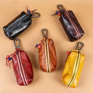 May include: Five leather key pouches in various colors: black, dark brown, red, tan, and yellow. Each pouch has a zipper closure, a metal clasp, and decorative beads. The pouches are designed to hold keys and small items.