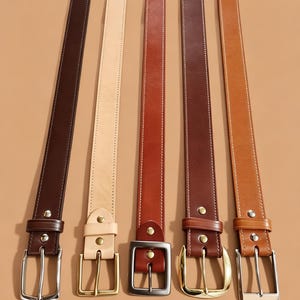 May include: Five leather belts in varying shades of brown and tan are displayed. Each belt has a different buckle style, including silver and gold-toned metal. The belts are arranged vertically, showcasing their length and color variations.