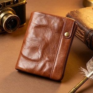 Puede incluir: Una cartera de cuero marrón con cierre de botón a presión. La cartera es rectangular y tiene un aspecto ligeramente desgastado. Una cámara vintage y un libro están en el fondo.