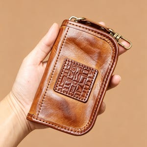 May include: A brown leather key case featuring a rectangular patch embossed with Egyptian hieroglyphics. The case has a smooth, polished finish with visible stitching along the edges and a brass-toned metal swivel clip attached to the top corner.