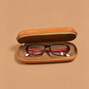 Könnte beinhalten: Eine Brille mit schwarzem Rahmen und roten Bügelinnenseiten, die in einem geöffneten, beigefarbenen, ovalen Hartschalenetui mit goldenem Reißverschluss und weichem, hellbraunem Innenfutter liegt.
