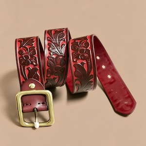 May include: A deep red leather belt featuring an intricate embossed floral pattern across the strap. The belt is equipped with a large, square-shaped brass-toned metal buckle and includes a series of circular adjustment holes along the end.