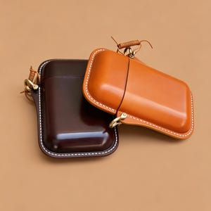May include: Two leather cases in shades of brown and tan. The cases have a rectangular shape with rounded edges, white stitching, and a small metal clasp. One case is a deep brown, the other is a lighter tan color.
