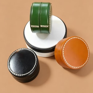 Puede incluir: Tres recipientes redondos con tapa en colores negro, verde y tostado. El recipiente verde se asienta sobre una plataforma circular blanca, mientras que los recipientes negro y tostado se sitúan debajo. Cada uno tiene costuras blancas.