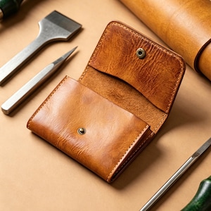 May include: A tan leather wallet with a visible grain texture, featuring a fold-over flap secured by a brass-colored metal snap button. The wallet is open, revealing an interior compartment, and is surrounded by various metal and wood leatherworking tools.