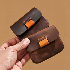 Puede incluir: Dos pequeñas carteras de cuero marrón oscuro con cierre de solapa y un acento de cuero naranja en contraste. Las carteras se sostienen en una mano sobre un fondo marrón. El cuero parece tener textura.