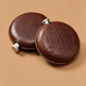 Peut inclure: Deux flasques rondes enveloppées de cuir marron foncé avec des bouchons à vis en métal argenté. La flasque avant présente des motifs gravés représentant une flèche, une ligne ondulée et une silhouette stylisée. Coutures claires sur les bords.
