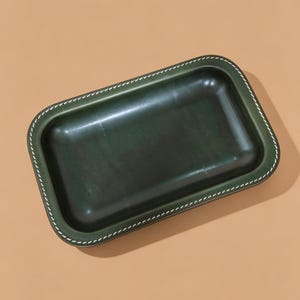 Puede incluir: Una bandeja rectangular de color verde oscuro con esquinas redondeadas y una superficie lisa y pulida. Los bordes de la bandeja están acentuados con un distintivo borde cosido en blanco que recorre todo el perímetro del borde elevado.