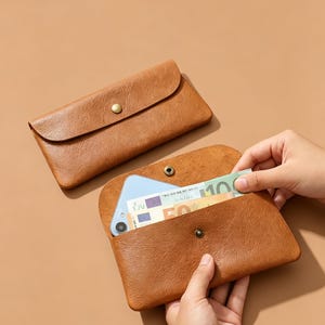 Op de afbeelding: Twee bruine leren portemonnees, één gesloten en één open, met een smartphone en valuta. De open portemonnee wordt vastgehouden. De portemonnees hebben een knoopsluiting.