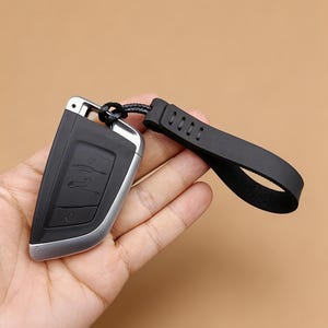 Puede incluir: Un mando de coche negro y plateado con un diseño elegante y angular que incluye botones integrados. El mando está unido a una correa de muñeca de silicona negra flexible mediante un cordón trenzado fino, facilitando su transporte seguro.