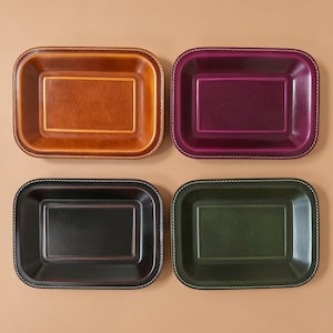 May include: Four rectangular trays with rounded corners, each featuring a smooth, polished surface and a stitched border. The set includes one tan, one deep purple, one black, and one dark green tray, all arranged in a two-by-two grid pattern.