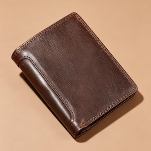 May include: A dark brown bi-fold wallet with a textured surface and visible stitching along the edges. The wallet features a smooth, rounded spine detail and a rectangular shape, presented in a closed position with a consistent deep brown tone.