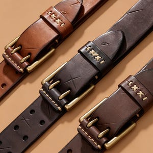 May include: Three leather belts in shades of tan, dark brown, and black. Each belt features a brass-toned metal buckle, decorative cross-stitch detailing in light thread, and etched X-shaped markings on the strap and the belt loop.